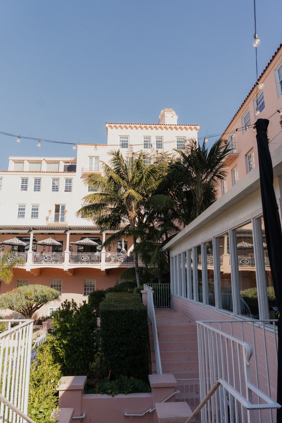 Pastel pink and white Mediterranean-style hotel building with palm trees and terraced walkways against blue sky.