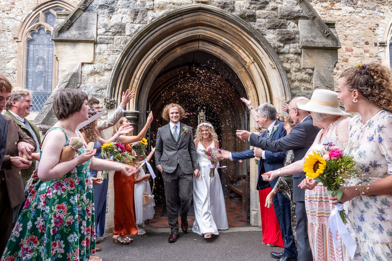 Wedding guests celebrate with confetti outside a stone church entrance archway during a joyous ceremony.