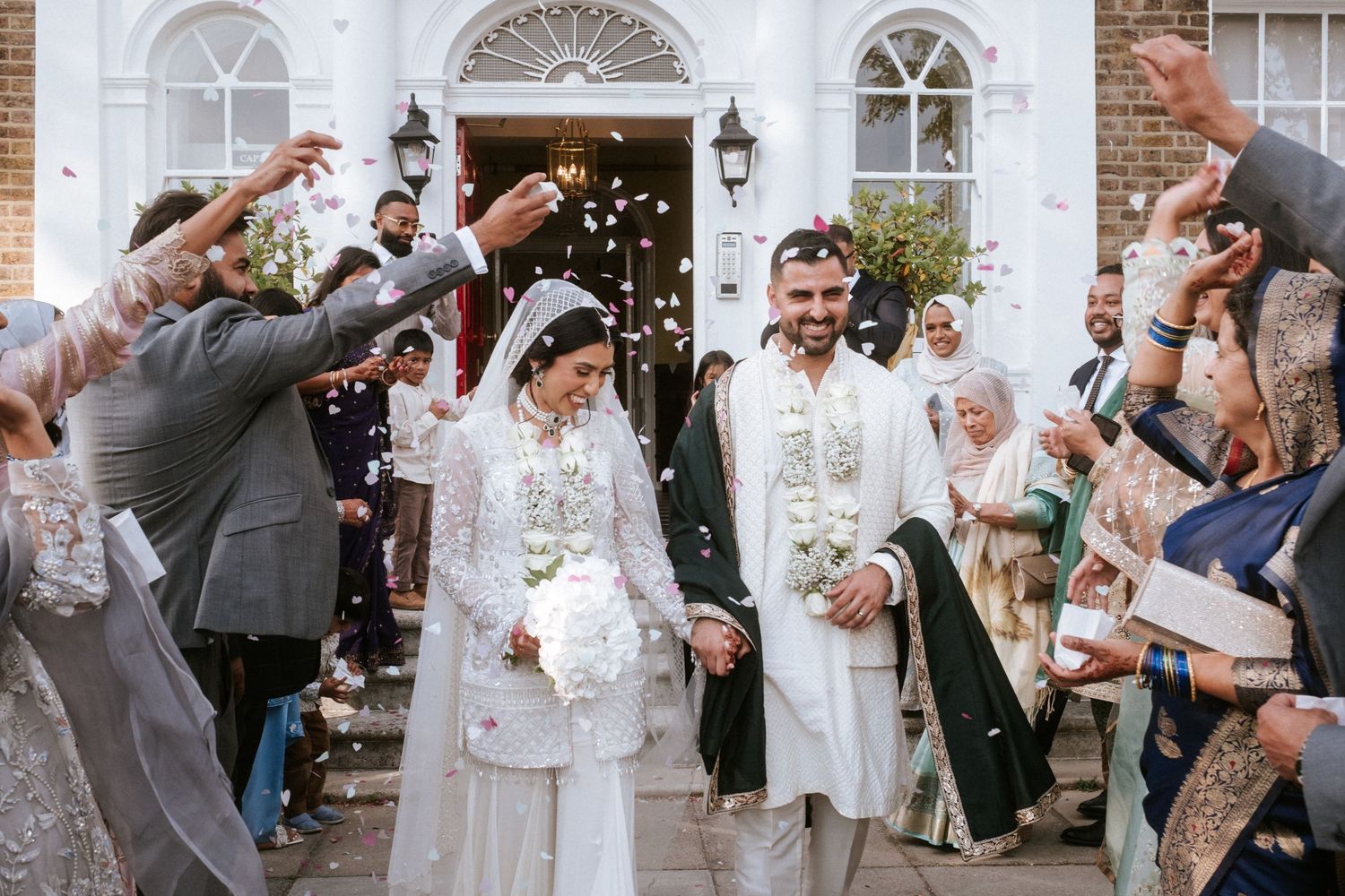 Wedding guests throw confetti and celebrate outside a historic brick building with arched windows.