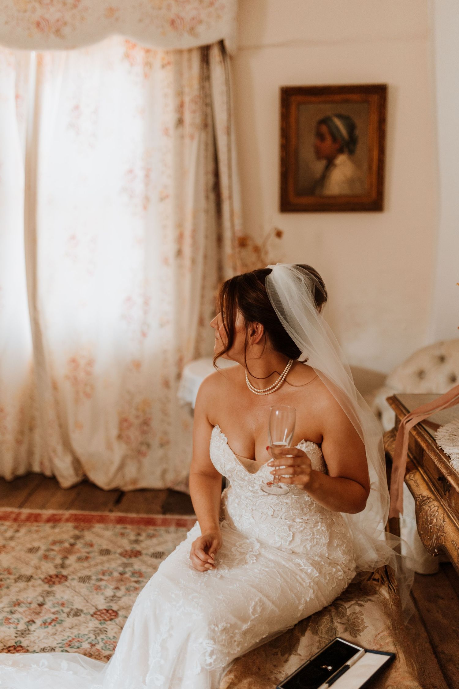 Bride in white lace wedding dress sits elegantly on vintage floral carpet in sunlit room with lace curtains at Lyde Court, Hereford.