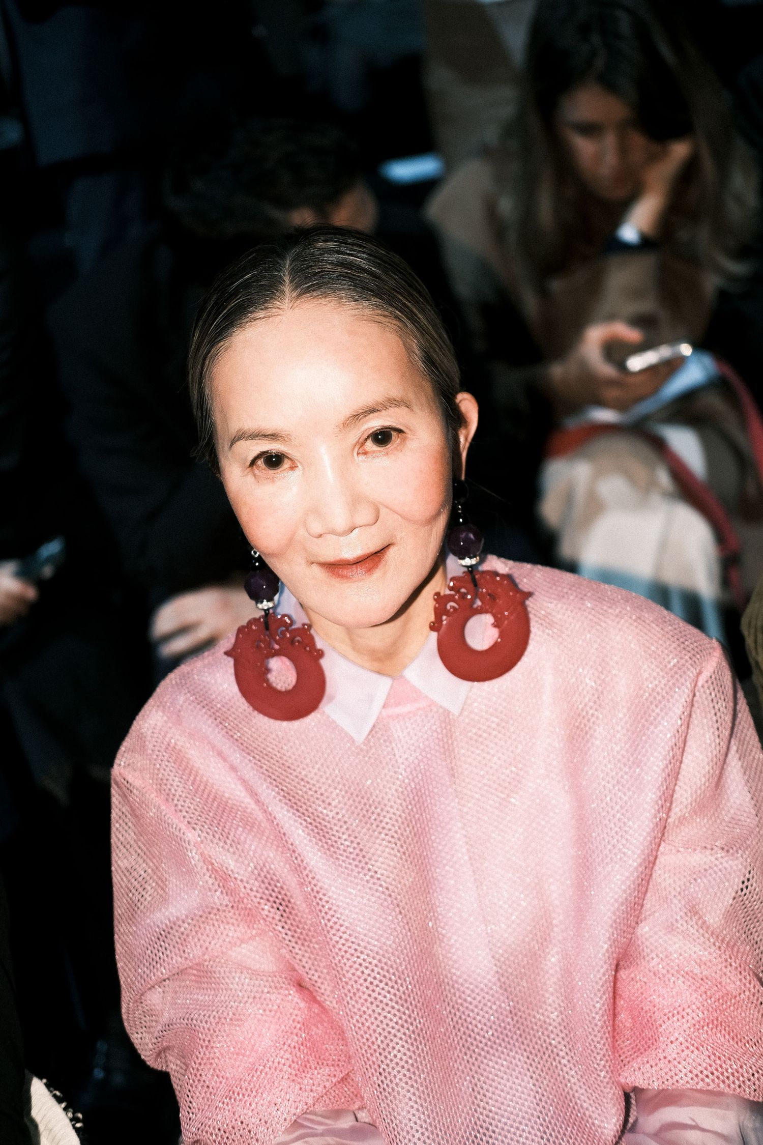 A person wearing a pink blouse smiles warmly in a dimly lit setting.