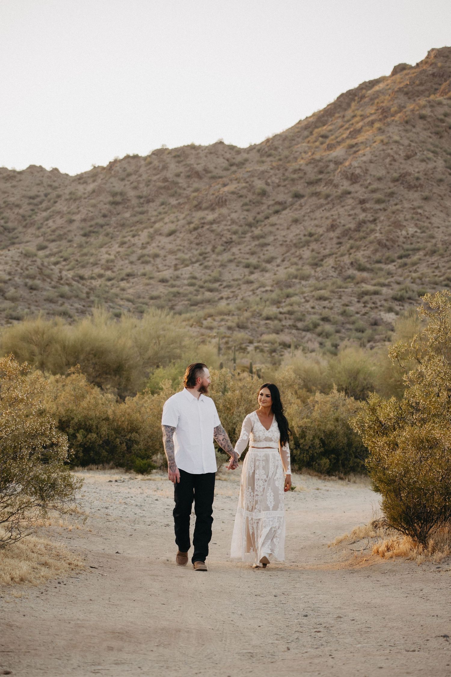 A couple walks hand in hand through a desert landscape at sunset with mountains in the background.