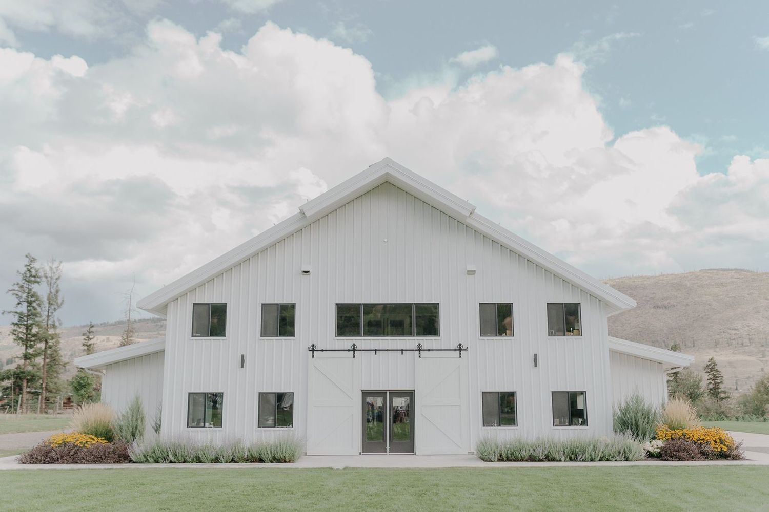 Large white barn wedding venue with sliding doors set against a mountain backdrop and cloudy sky.