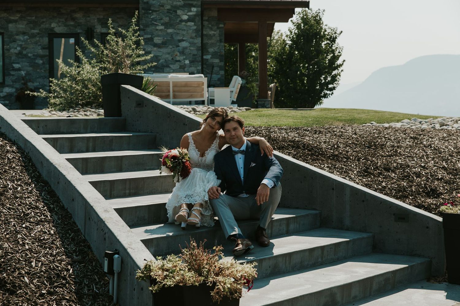Bride and groom sitting together on stone steps outside rustic venue surrounded by lush greenery.