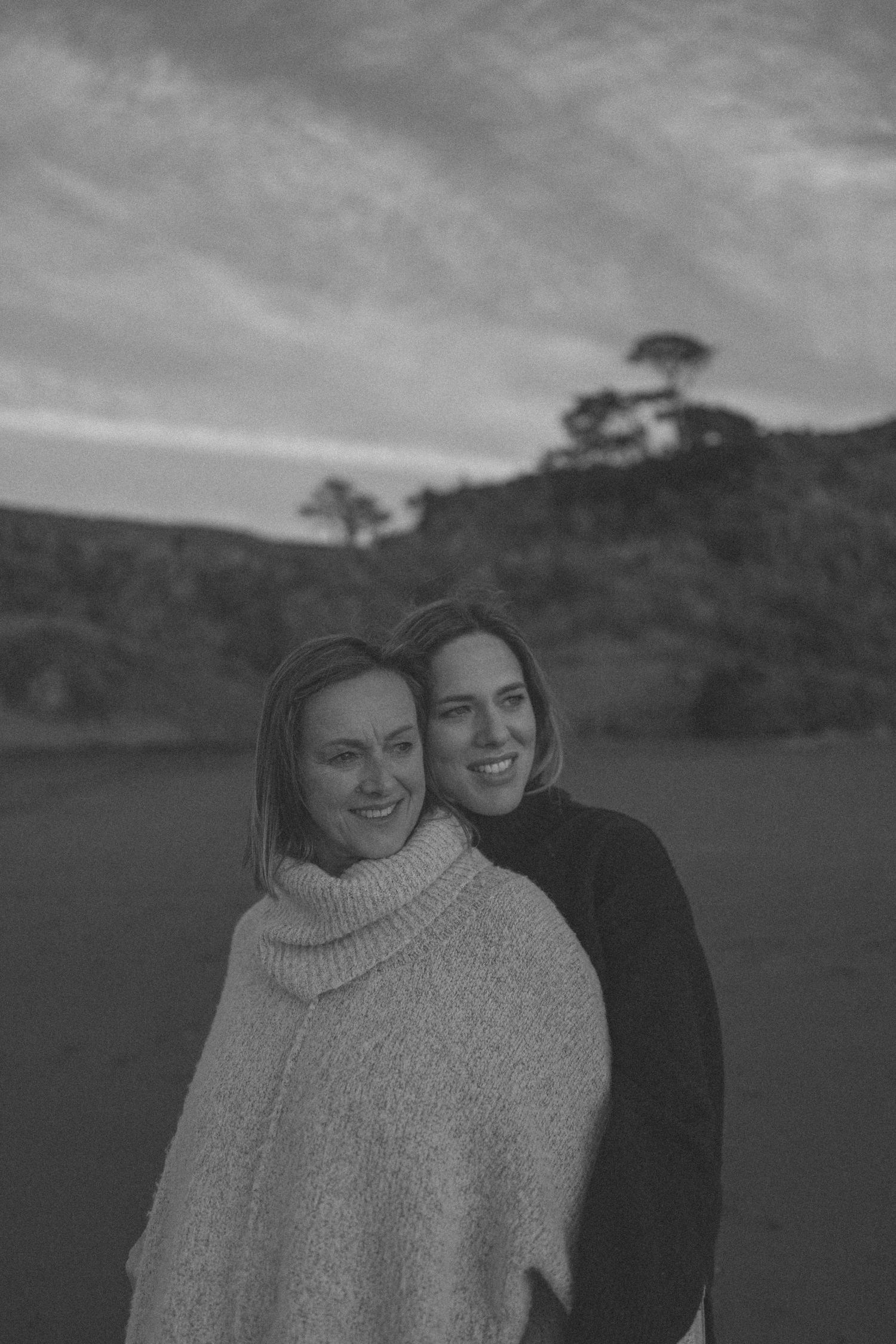 Black and white portrait of two smiling women standing together at dusk with hills in the background in New Zealand.