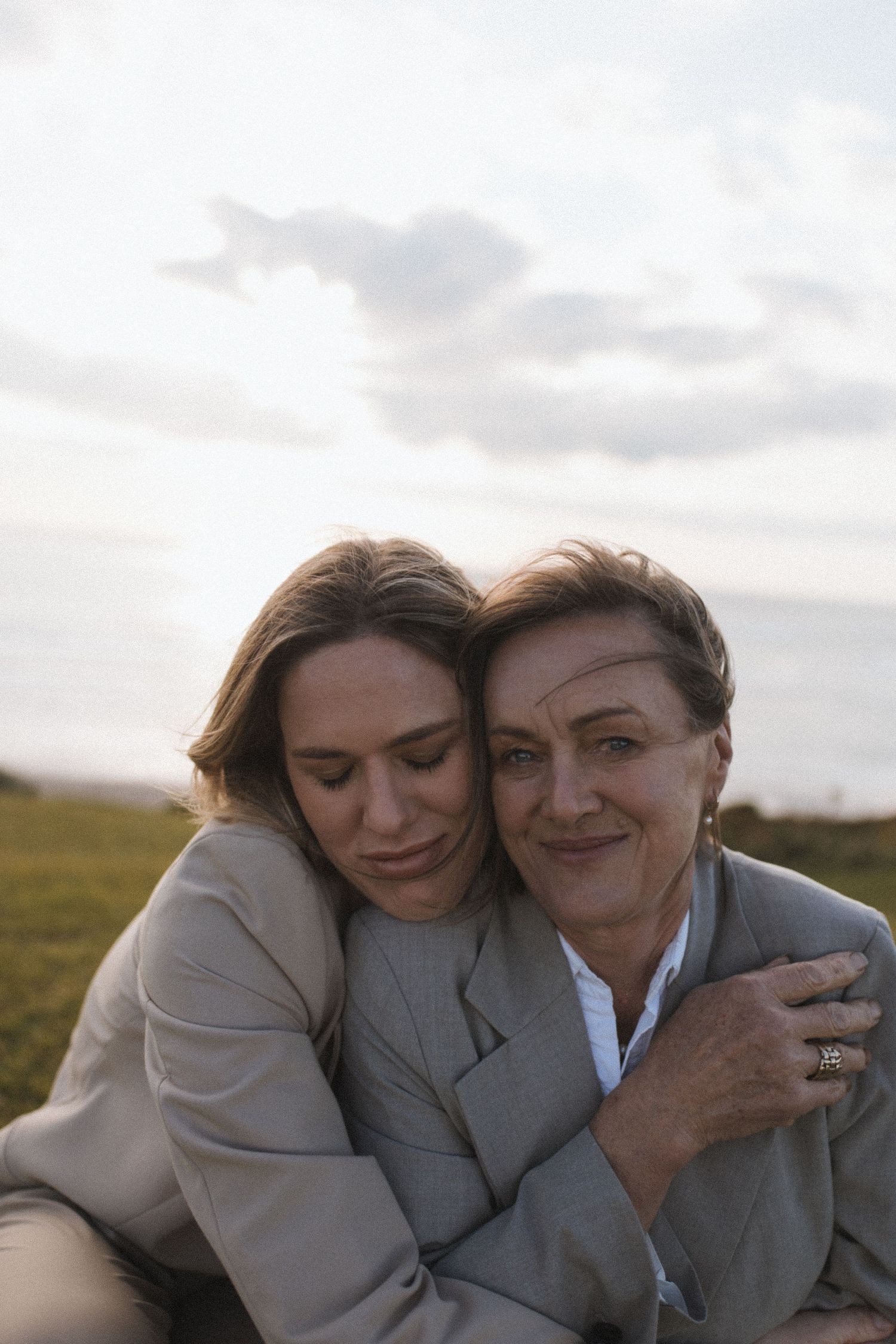 A young woman embraces an older woman outdoors in a green field under a cloudy sky, both wearing gray blazers in New Zealand.