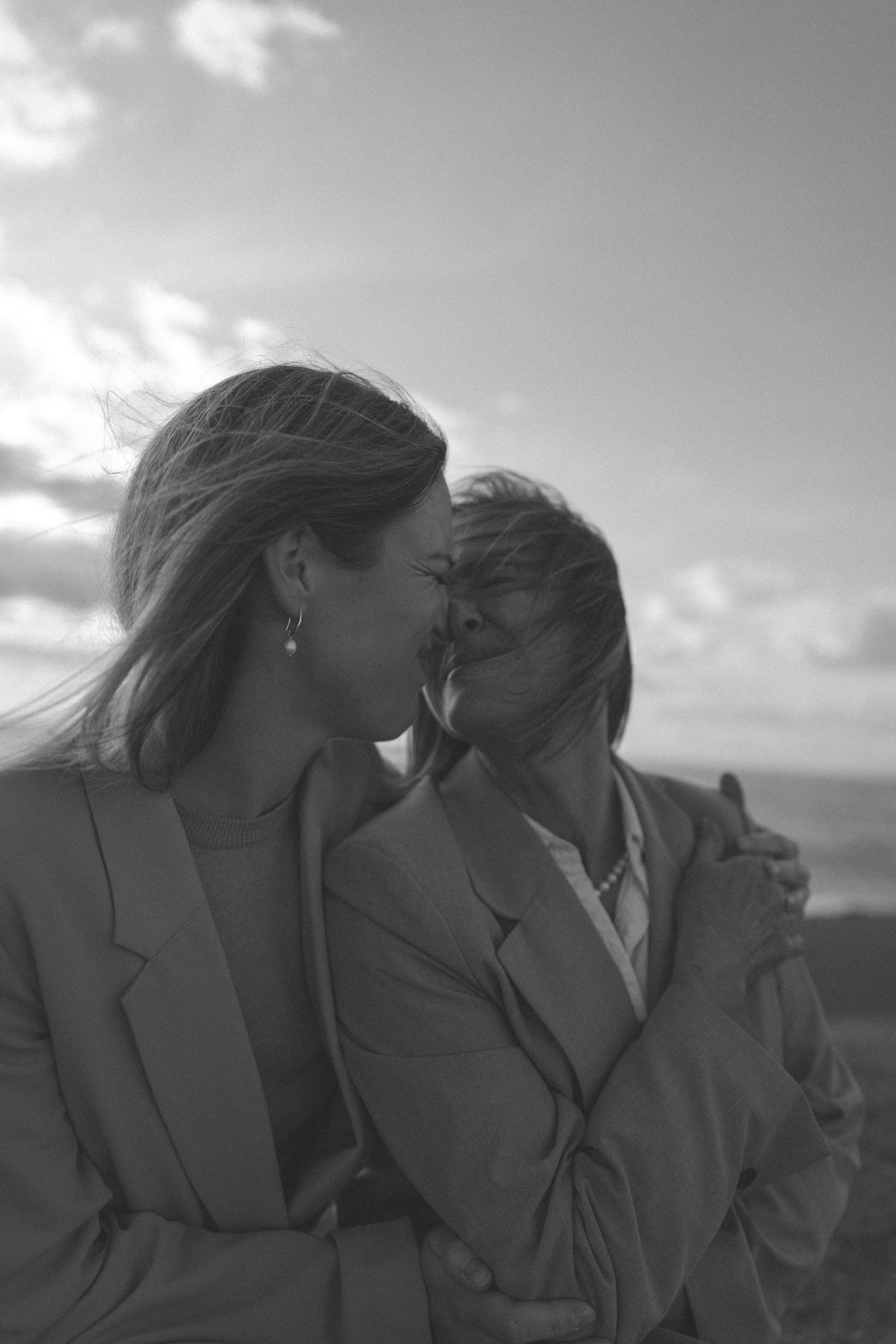 Black and white photo of two women in blazers sharing a tender moment outdoors under a cloudy sky in New Zealand.