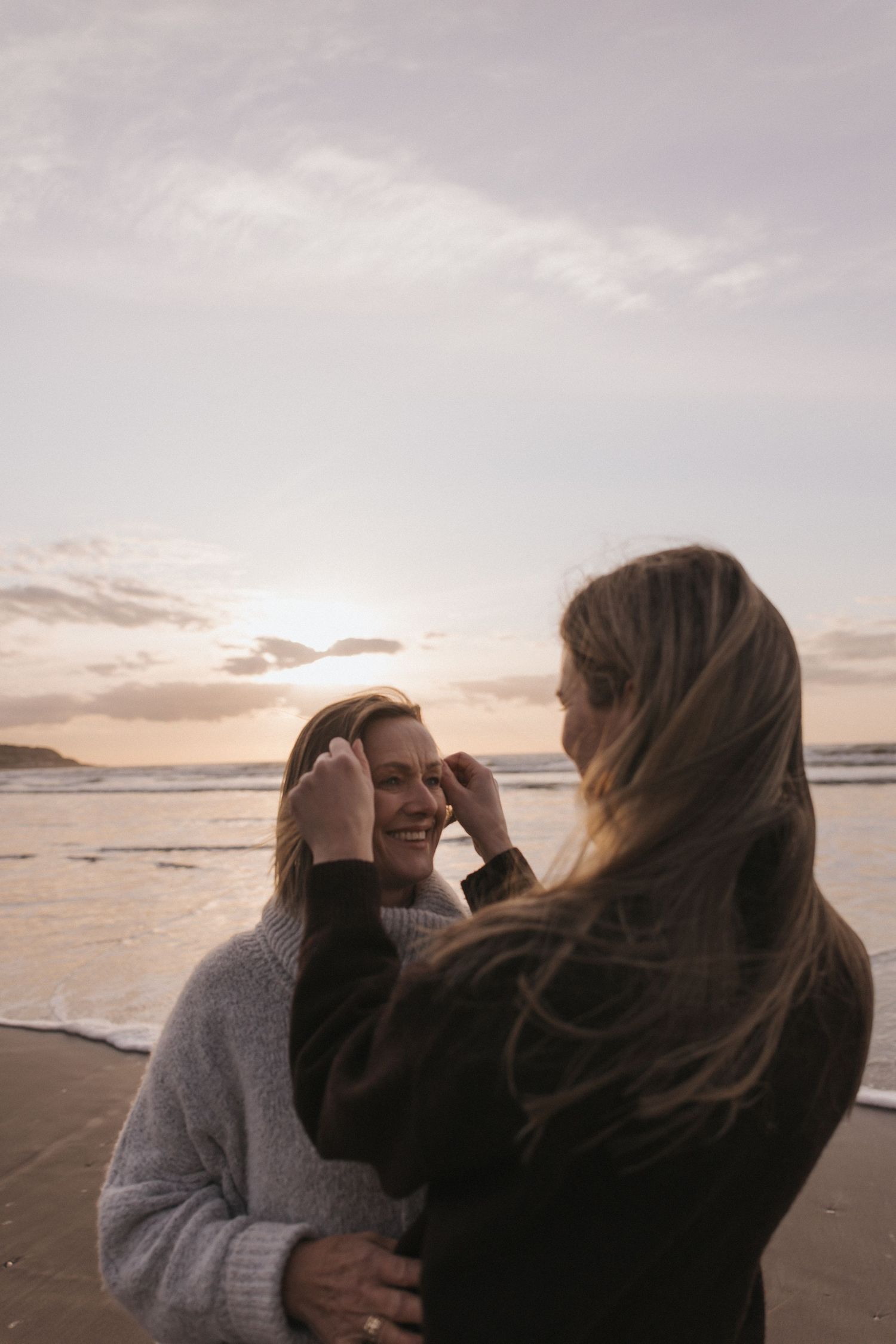 Two women embrace on a sandy beach at sunset, one tucking hair behind the others ear in New Zealand.