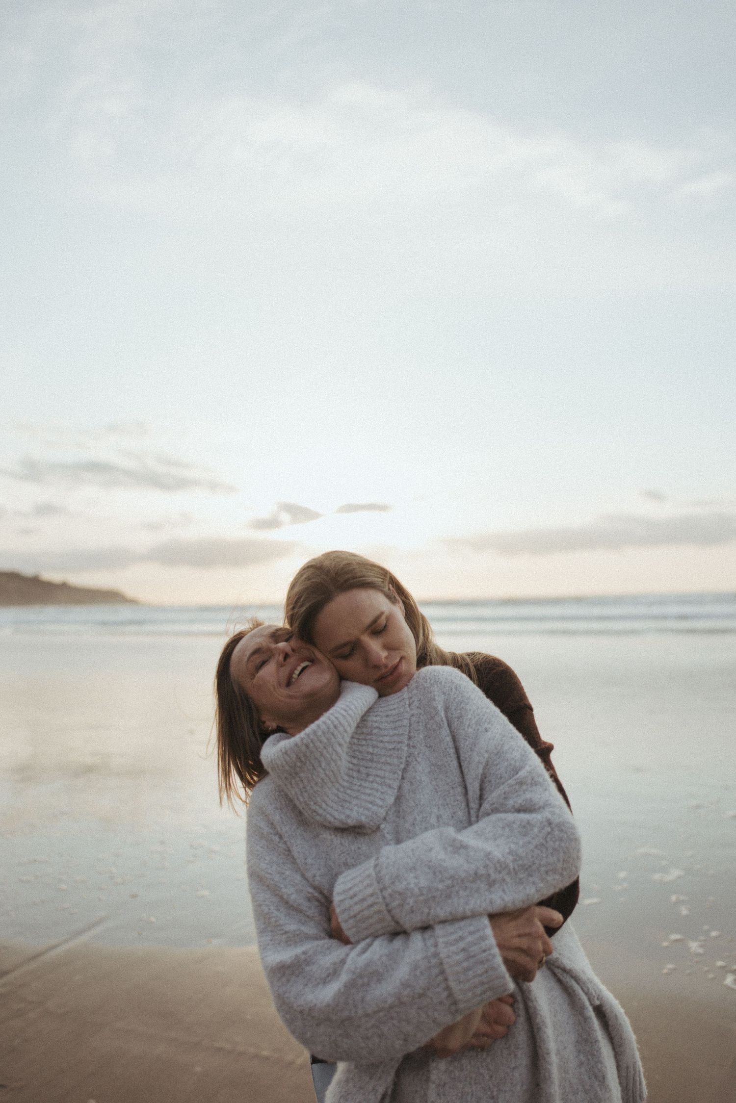 Two women share a warm embrace on a beach at sunset, wrapped in cozy knit sweaters in New Zealand.