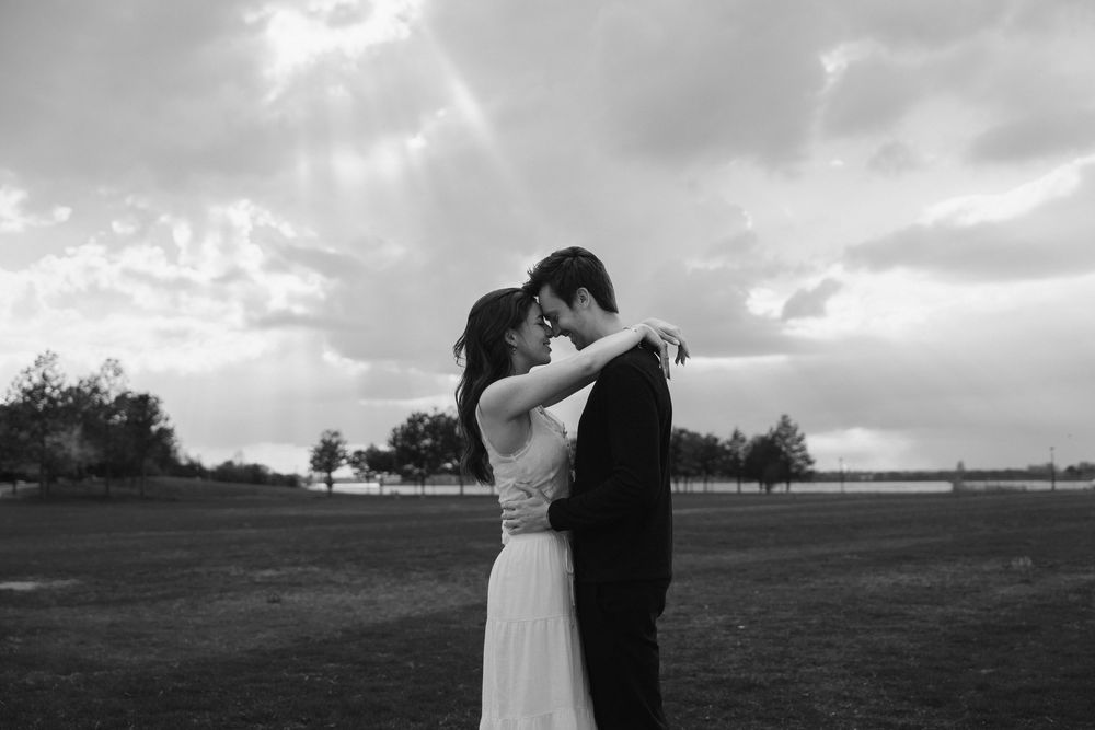 A black and white portrait of an embrace in an open field under dramatic clouds.