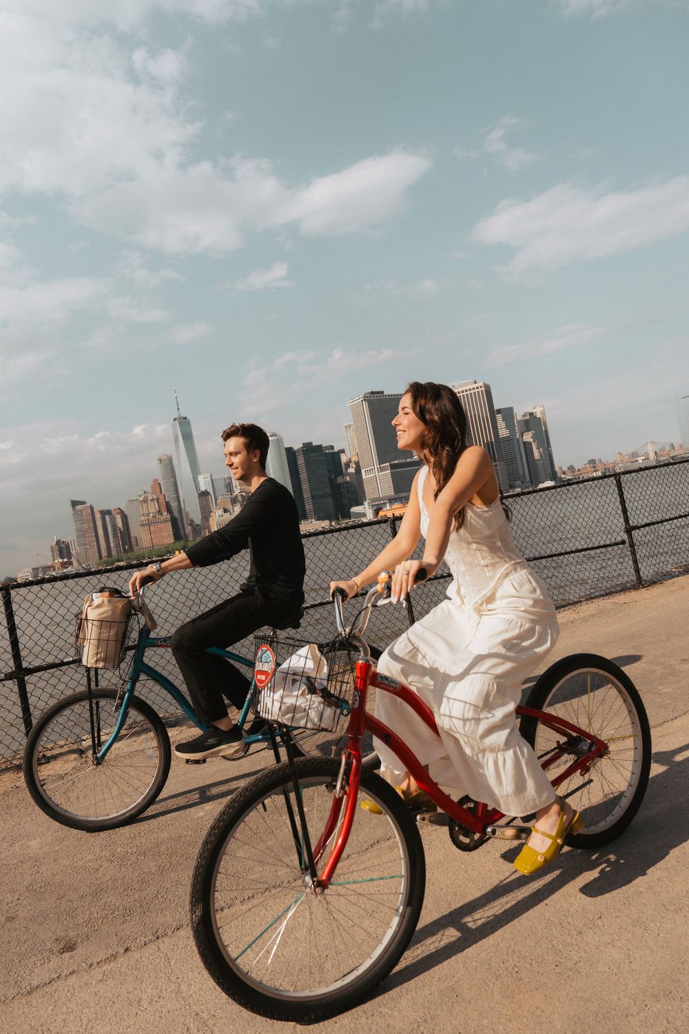A couple rides bicycles along a waterfront path with the Manhattan skyline visible in the background.