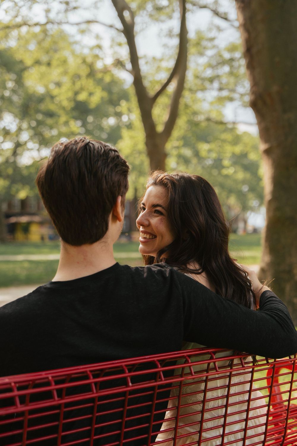 A couple shares an intimate moment while sitting on a red bench in a sunlit park.