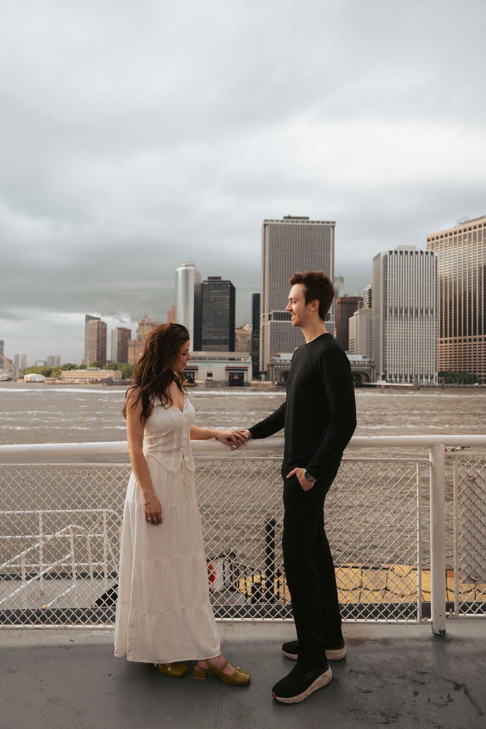 A couple shares a moment on a ferry deck with the NYC skyline visible behind them.