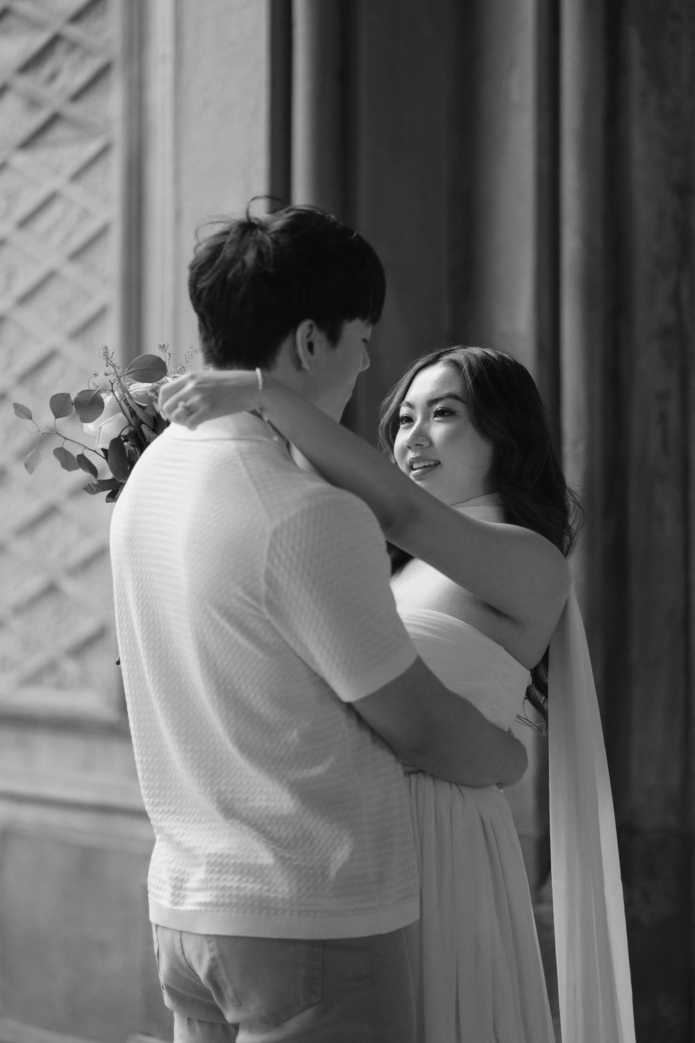 A romantic couple embraces in an intimate black and white portrait against an architectural backdrop.