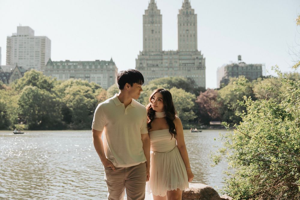 A couple poses by the lake in Central Park with the iconic Manhattan skyline behind them.