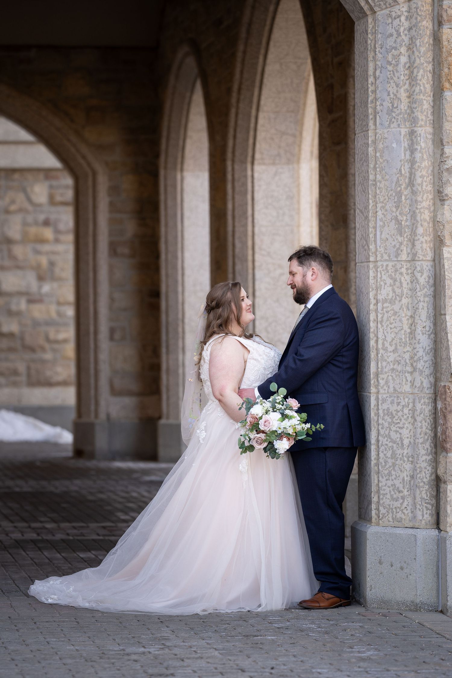 A bride and groom share a tender moment under stone archways while holding a white and blush floral bouquet.