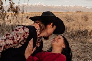 A couple in western attire shares a romantic moment in a rustic field with mountains visible in the background.