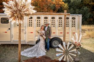 Wedding couple poses in front of vintage livestock trailer decorated with pampas grass and wagon wheels.