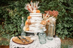 Elegant wedding cake display on lace tablecloth with rustic decorative elements and greenery in background.