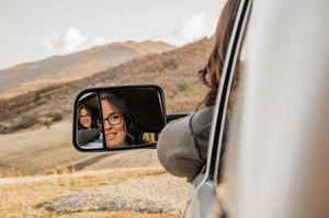 Side mirror reflection view of silver truck driving through mountain landscape.