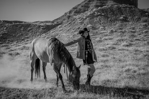 A black and white scene of a horse grazing while someone in western wear stands nearby.