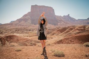 A figure in a black dress strikes a pose against towering red rock formations in the desert.