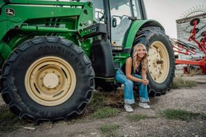 Someone in denim sits beside the massive wheel of a John Deere tractor.