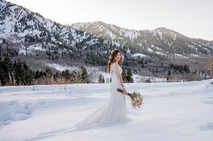Bride in flowing white gown stands in winter mountain landscape with snow.