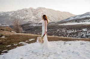 Elegant bride in white dress poses in snowy mountain landscape with bouquet.