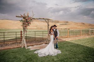 Wedding ceremony setup with rustic wooden arch and white fabric against rolling hills and cloudy sky.