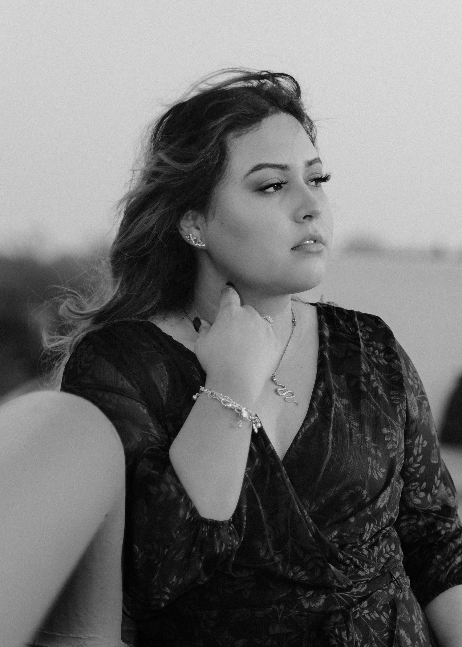 Black and white portrait of a woman in a dark wrap dress with jewelry looking to the side in soft natural lighting.