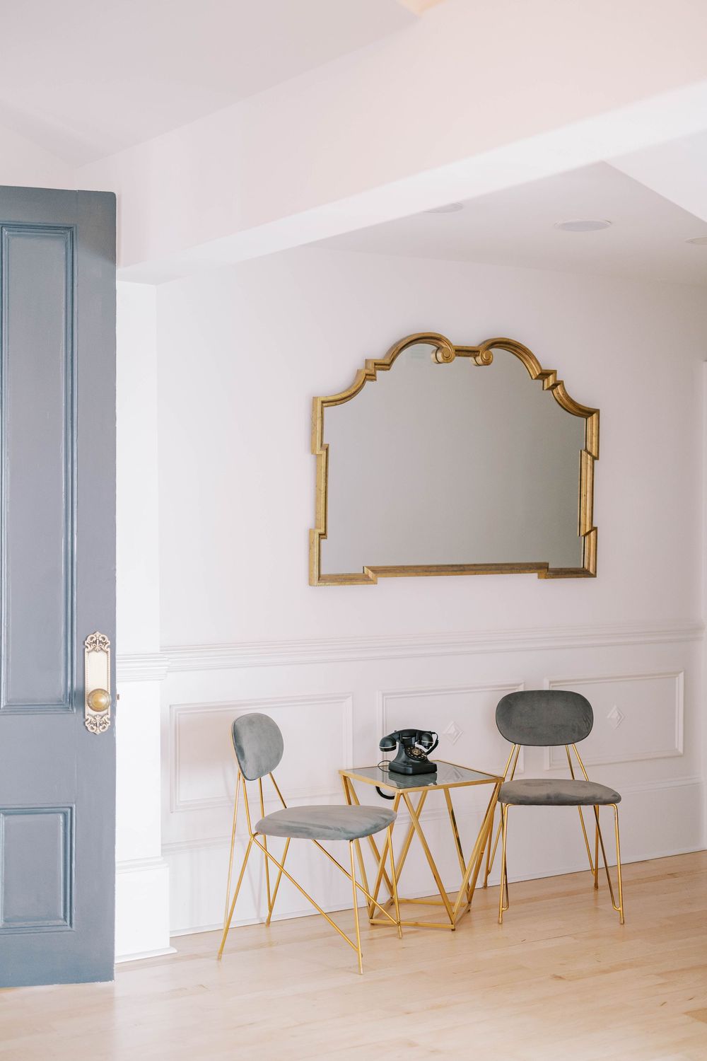 Modern seating area with gold-framed mirror, gray chairs and round table against white paneled wall.