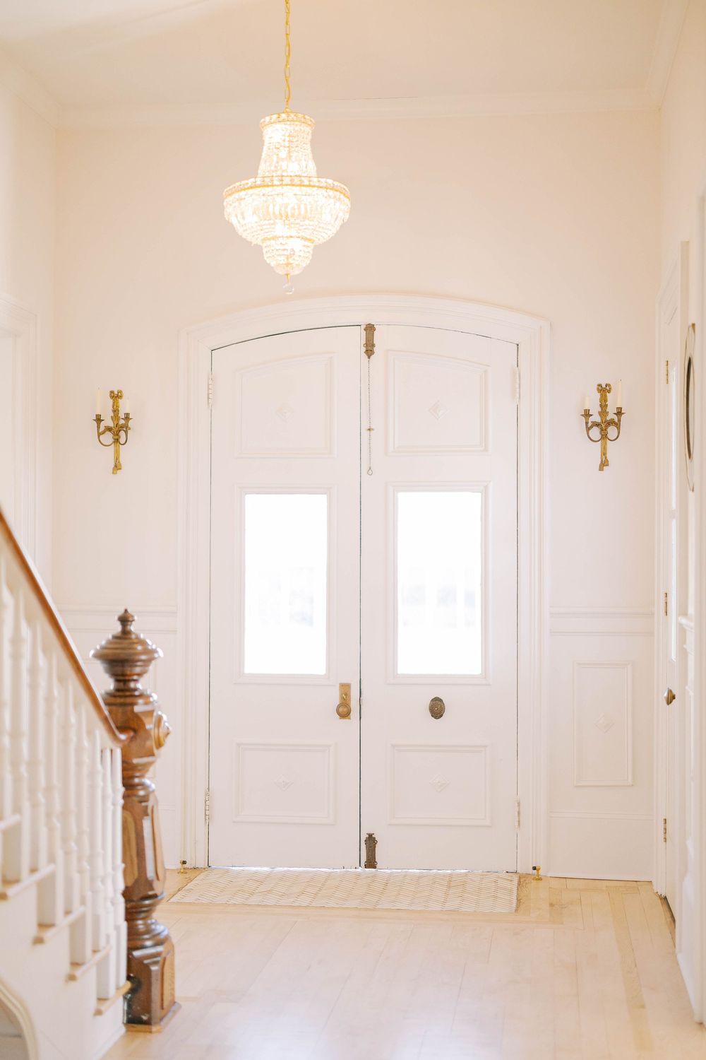Elegant white entryway with crystal chandelier, wooden banister, and double front doors.