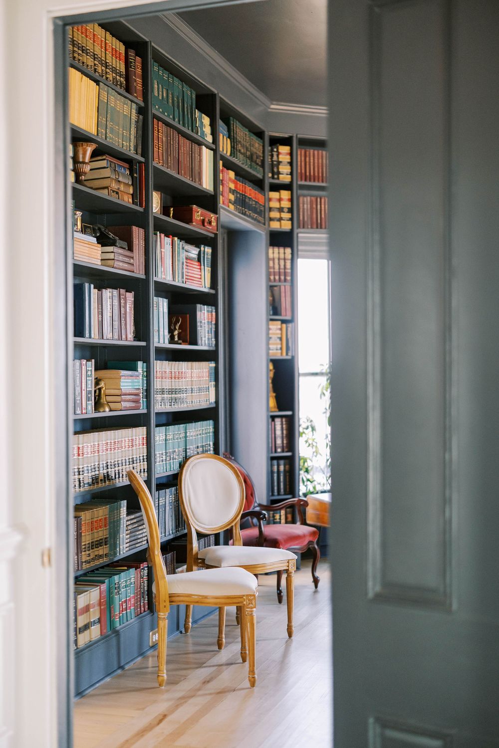Elegant library hallway with dark built-in bookshelves and antique chairs along a bright window-lit corridor.