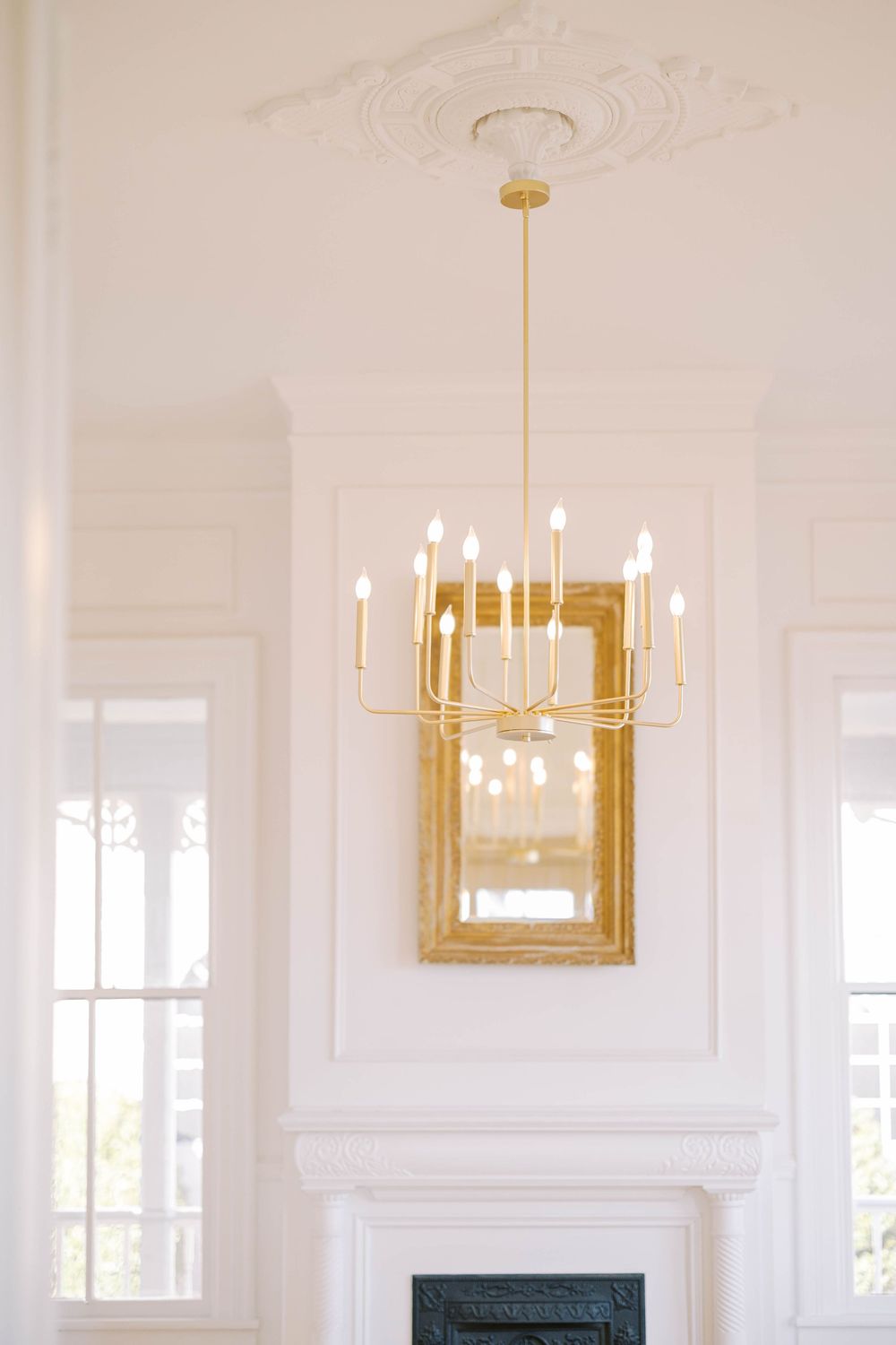 Elegant white room with gold chandeliers, crown molding, and large windows creating a bright luxurious space.