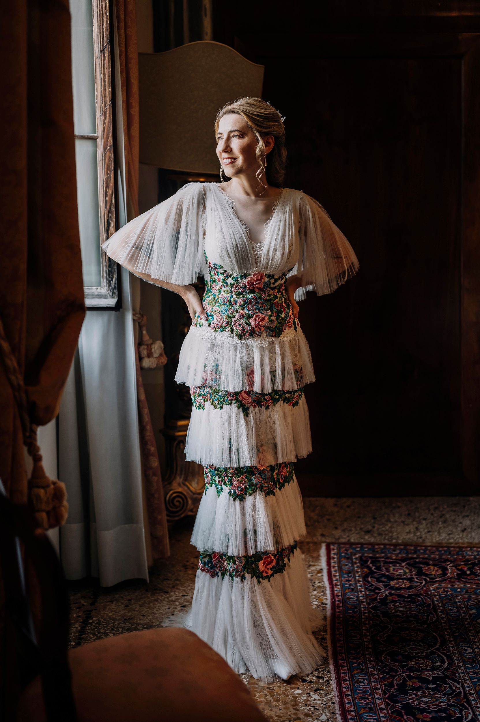 Sposa che indossa un abito bianco a balze di ispirazione vintage con decorazioni floreali in un ambiente interno buio.