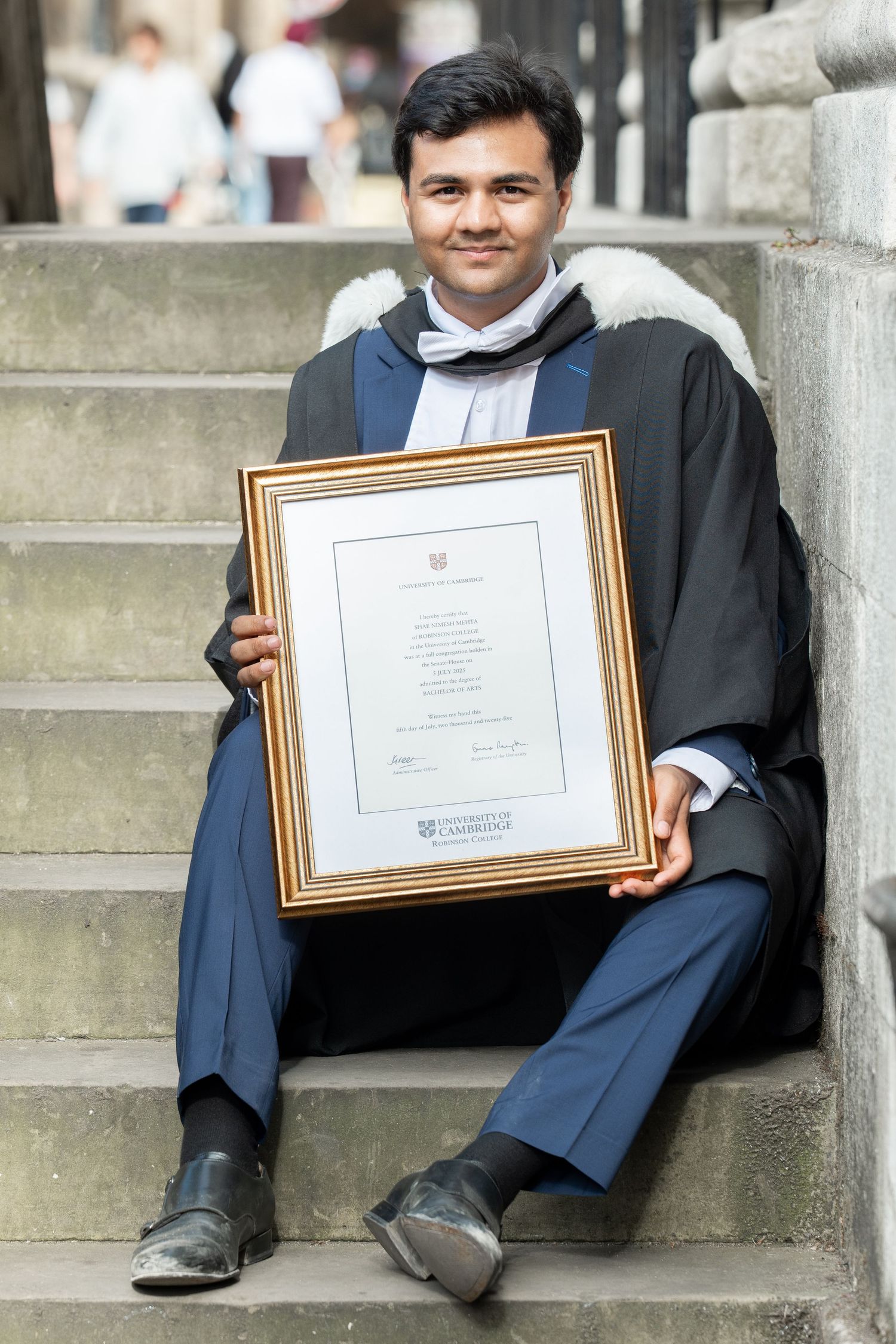 A graduate in academic regalia sits on stone steps while holding a framed diploma certificate.