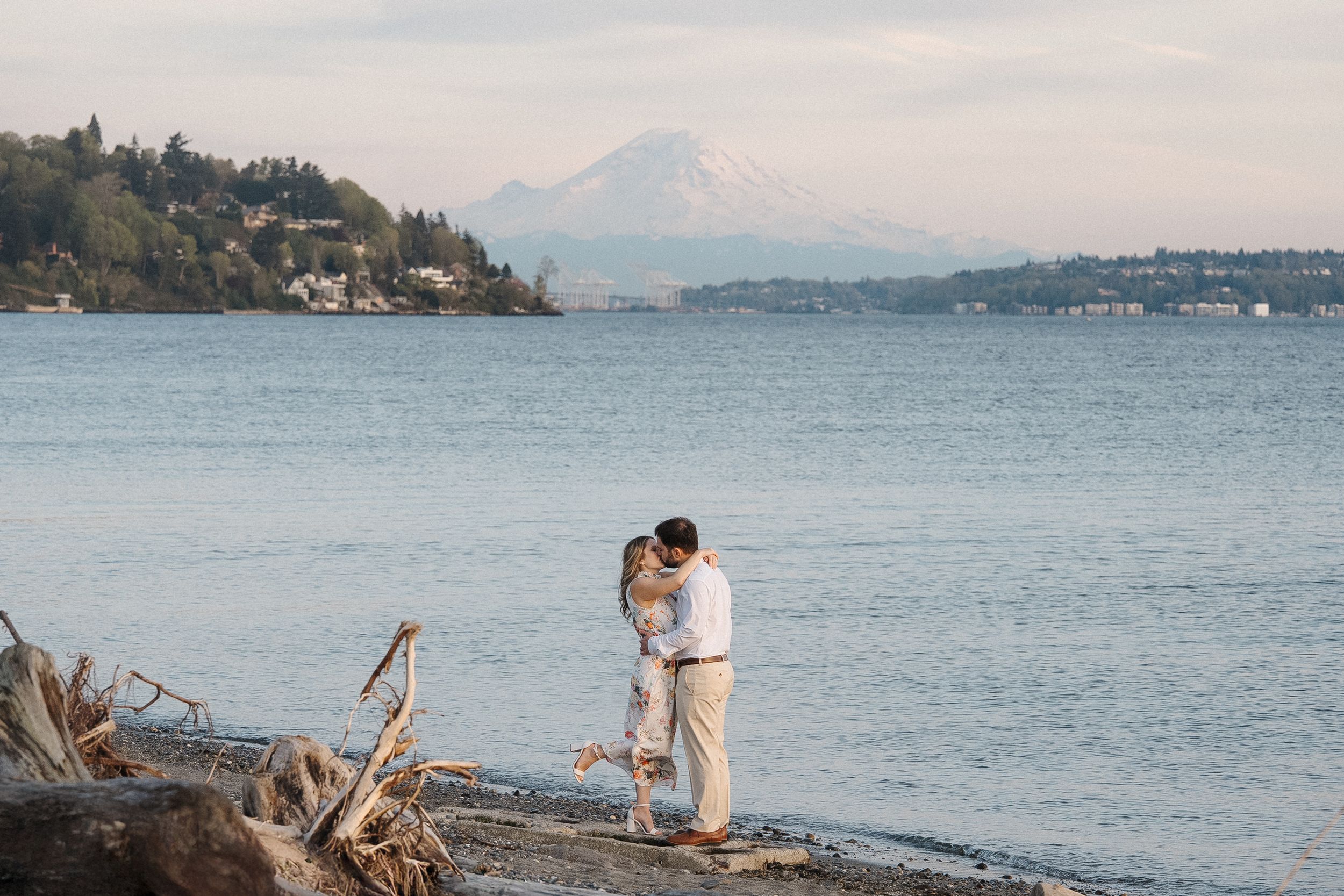 Romantic couple embraces on a rocky beach overlooking calm blue waters and distant mountains at sunset.