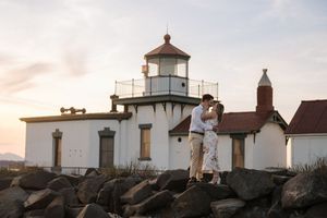 West Point Lighthouse in Seattle stands against a cloudy sky with its white tower and brown trim at sunset.