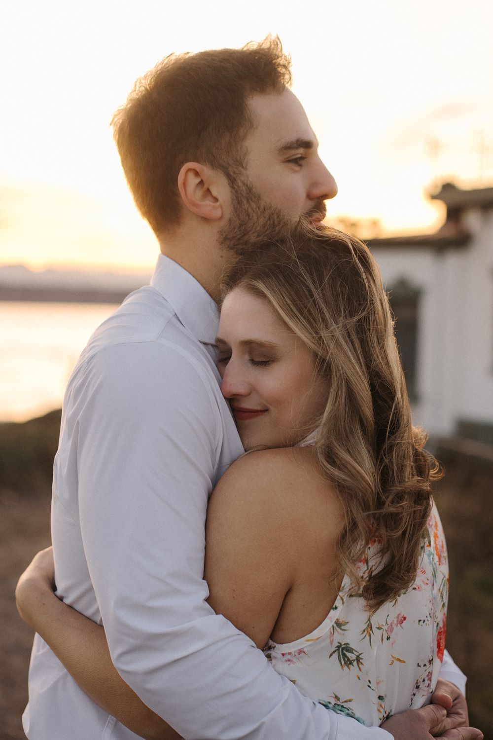A couple shares an intimate embrace during golden hour, with the person wearing a white shirt hugging their partner in a floral dress.