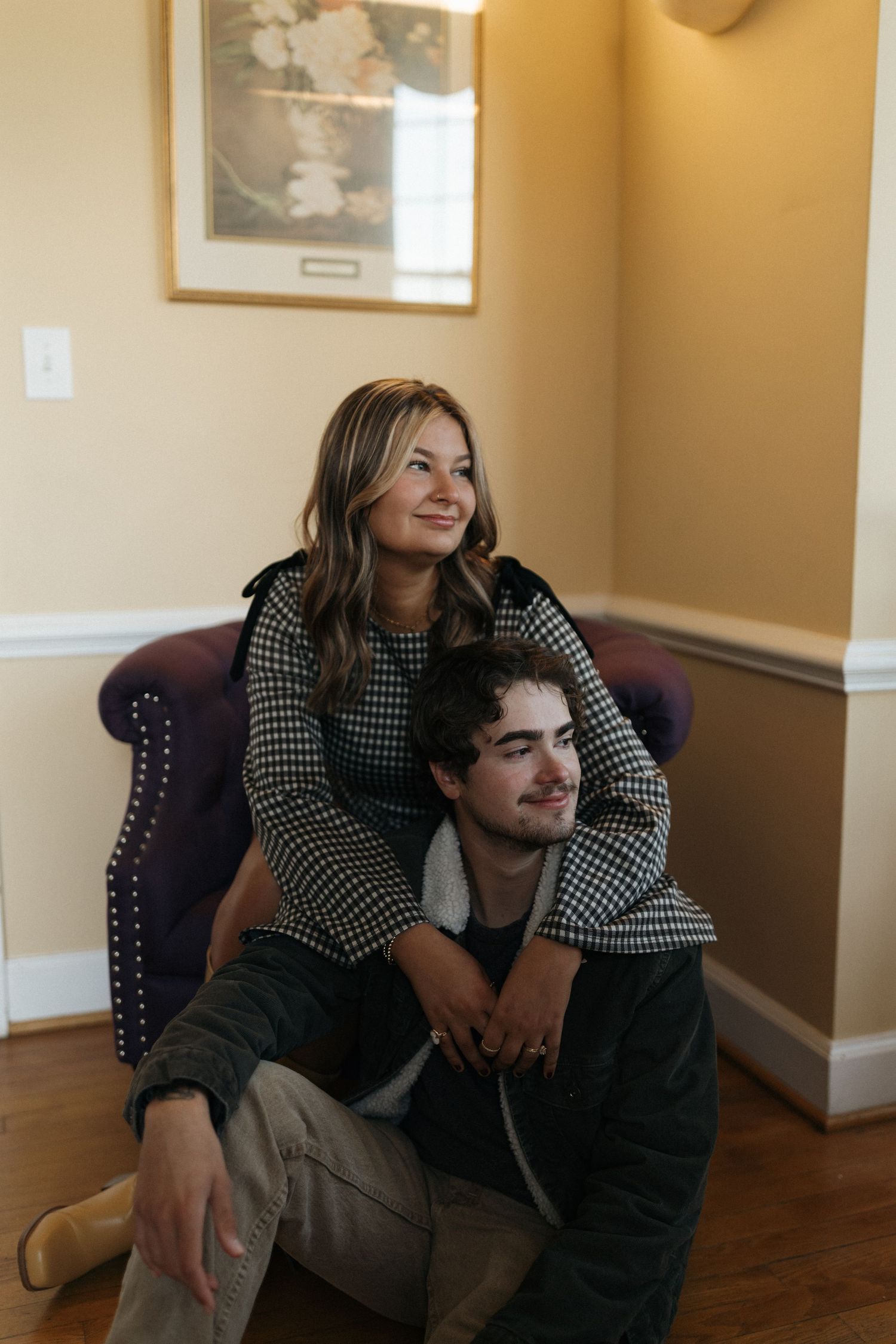 Two people share an intimate moment sitting on the floor in a warmly lit room with hardwood floors and beige walls.