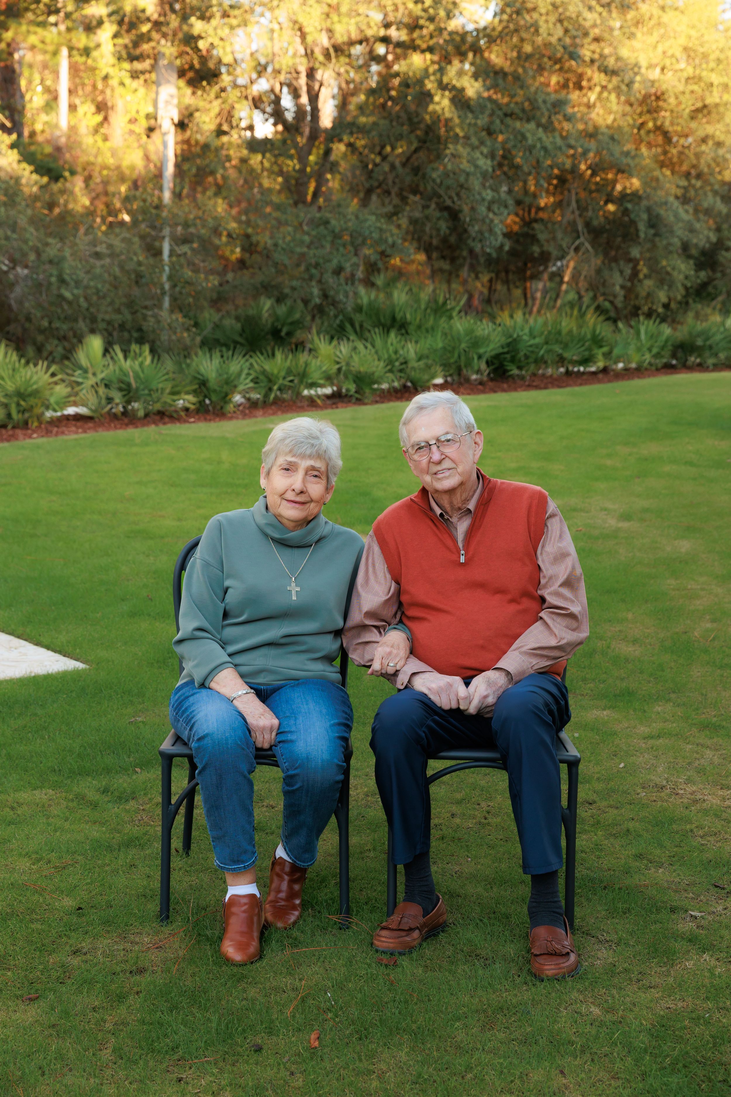 Two seniors sit together on a bench in a sunny garden wearing casual clothes and sharing a sweet moment.