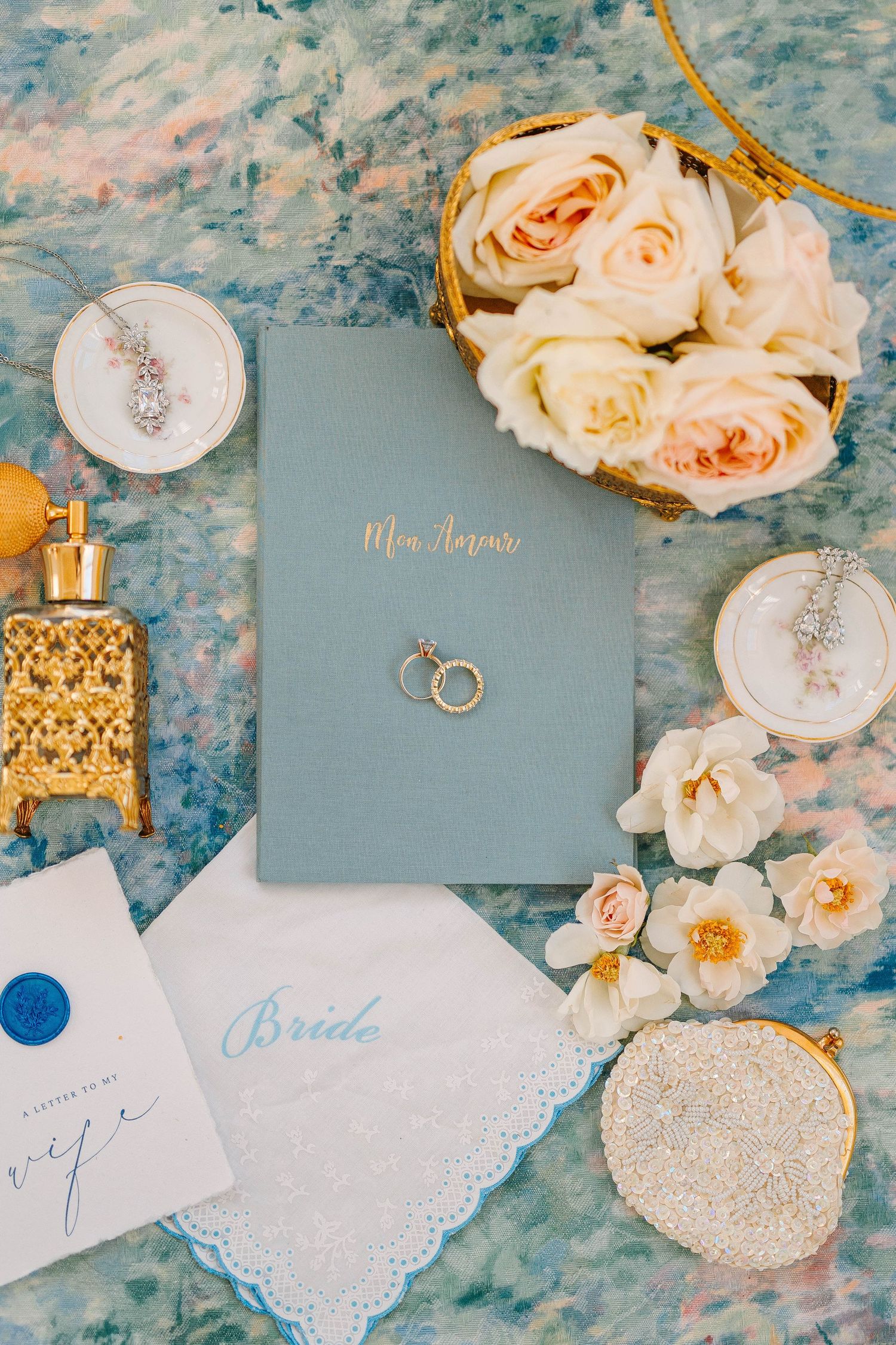 Wedding details flatlay with pale pink roses, blue vow book, rings, and bridal accessories on marbled surface.