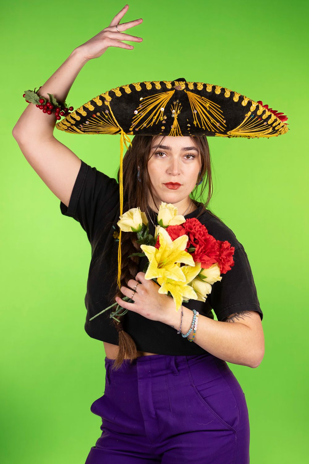 Person in black shirt wearing decorative sombrero holding colorful flowers against bright green background.
