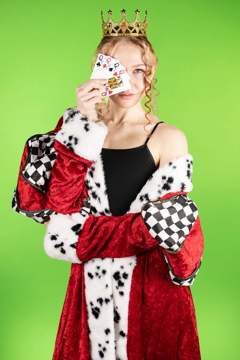 Person in royal costume with crown and dalmatian print robe holds playing cards against green background.