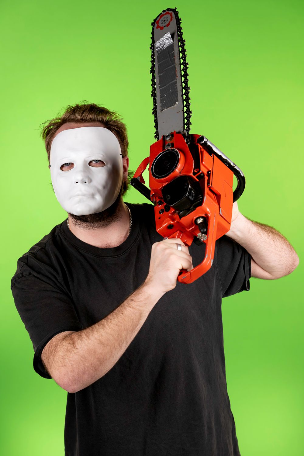 Person in white mask and black shirt poses with chainsaw against green backdrop.