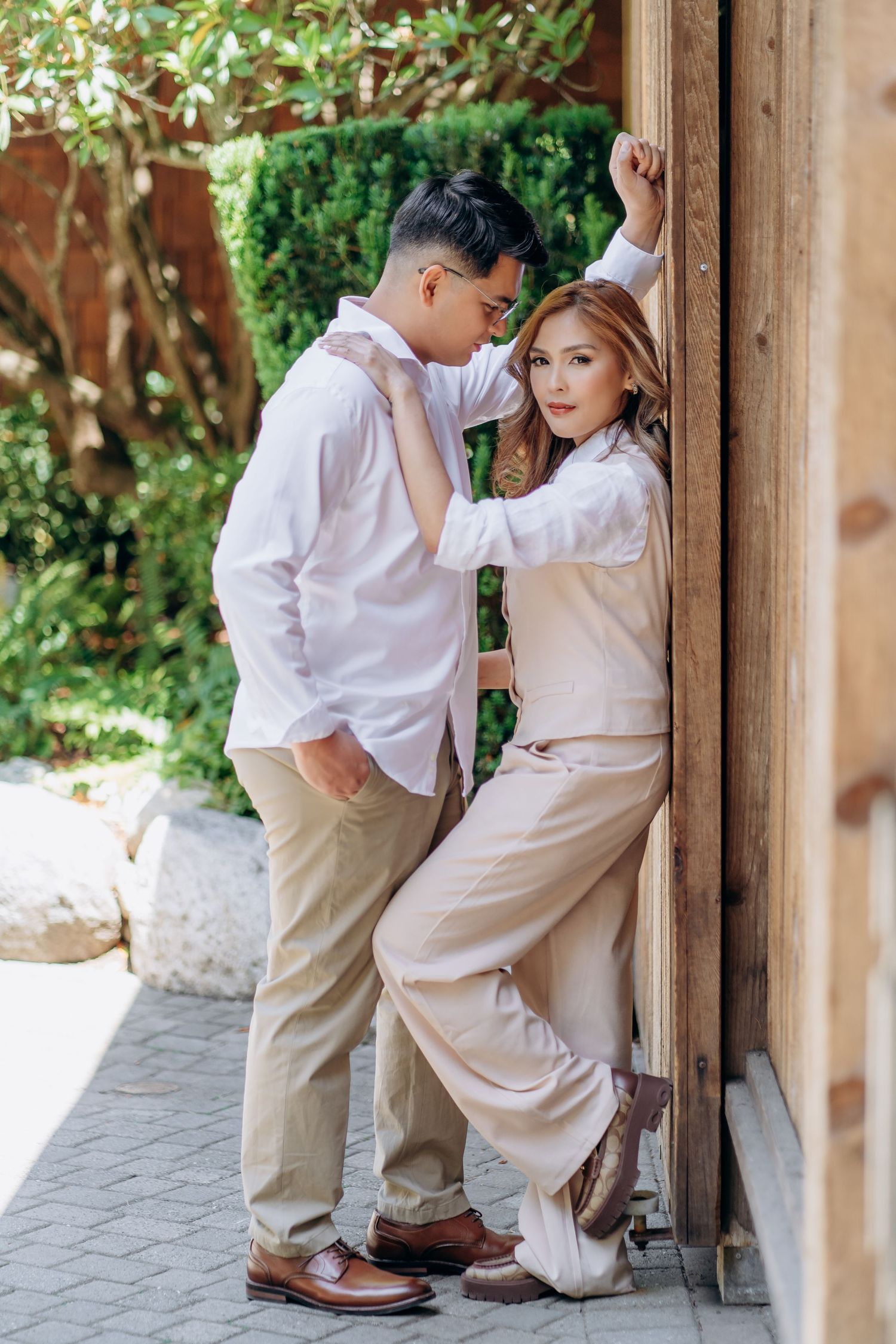 A romantic vintage-style photoshoot features a couple in light colored outfits posing against wooden pillars outdoors.