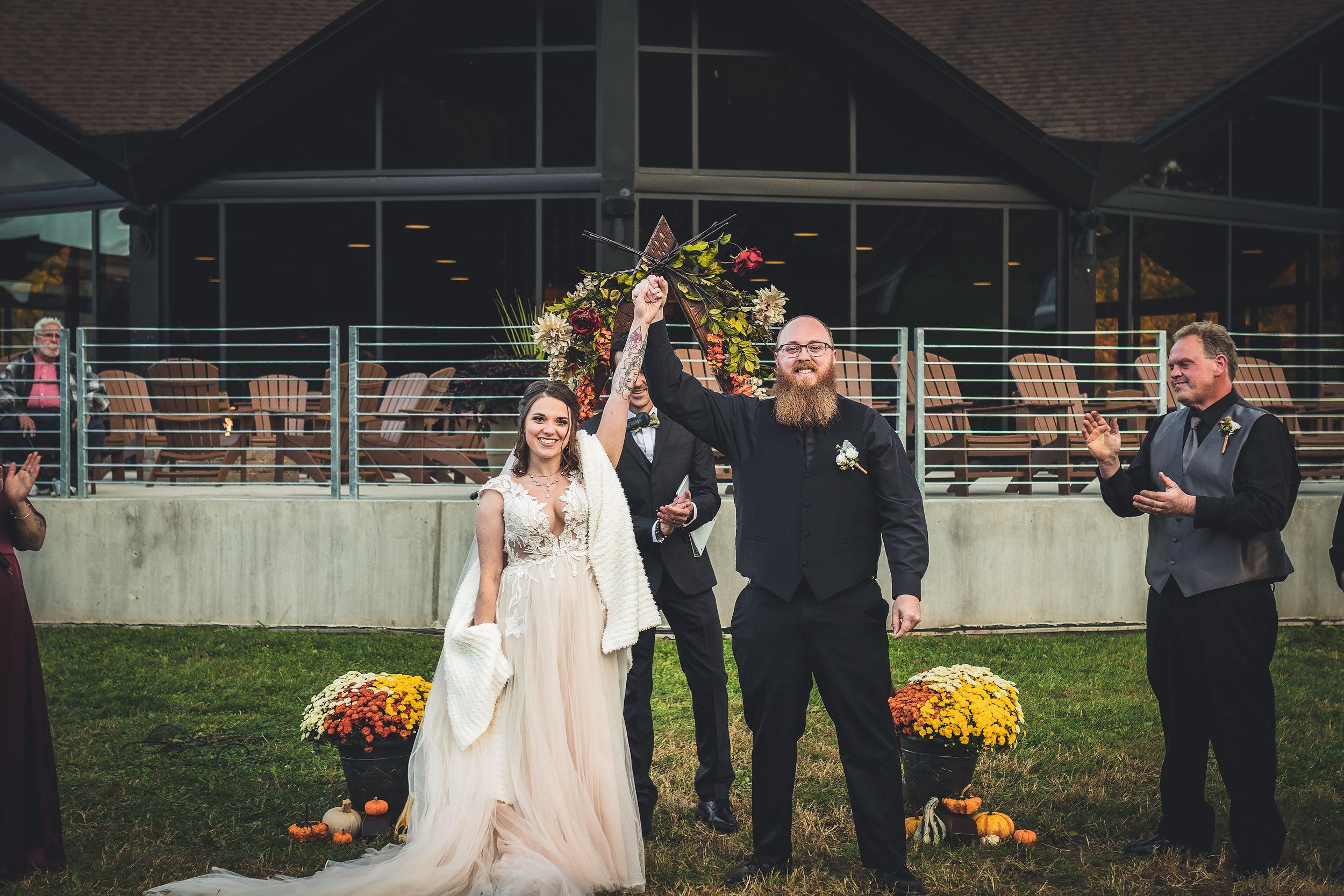 Wedding ceremony guests gather outdoors with yellow floral decorations and autumn foliage in the background.