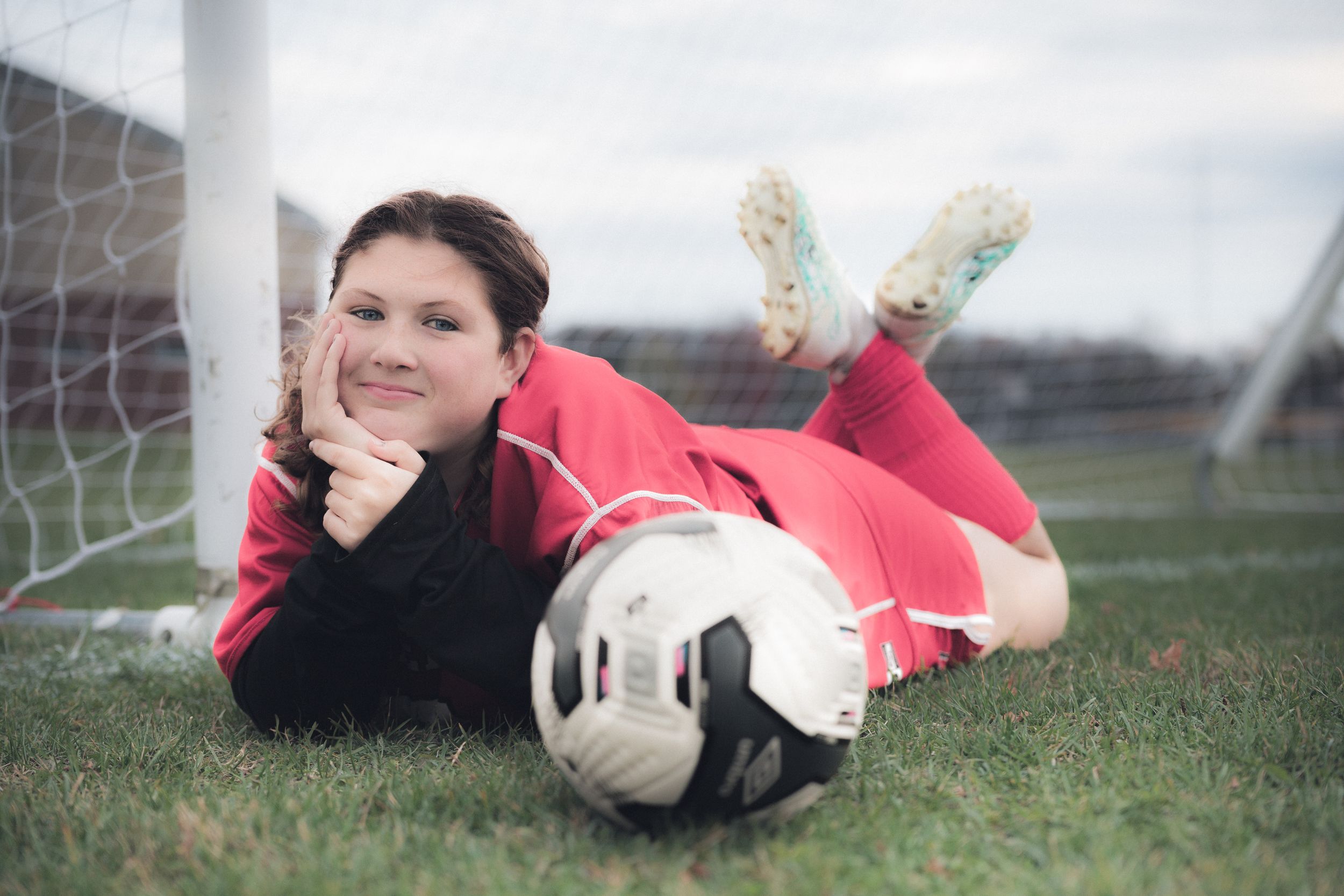 Soccer-themed photo shoot featuring a goalkeeper lying on grass field in red uniform.