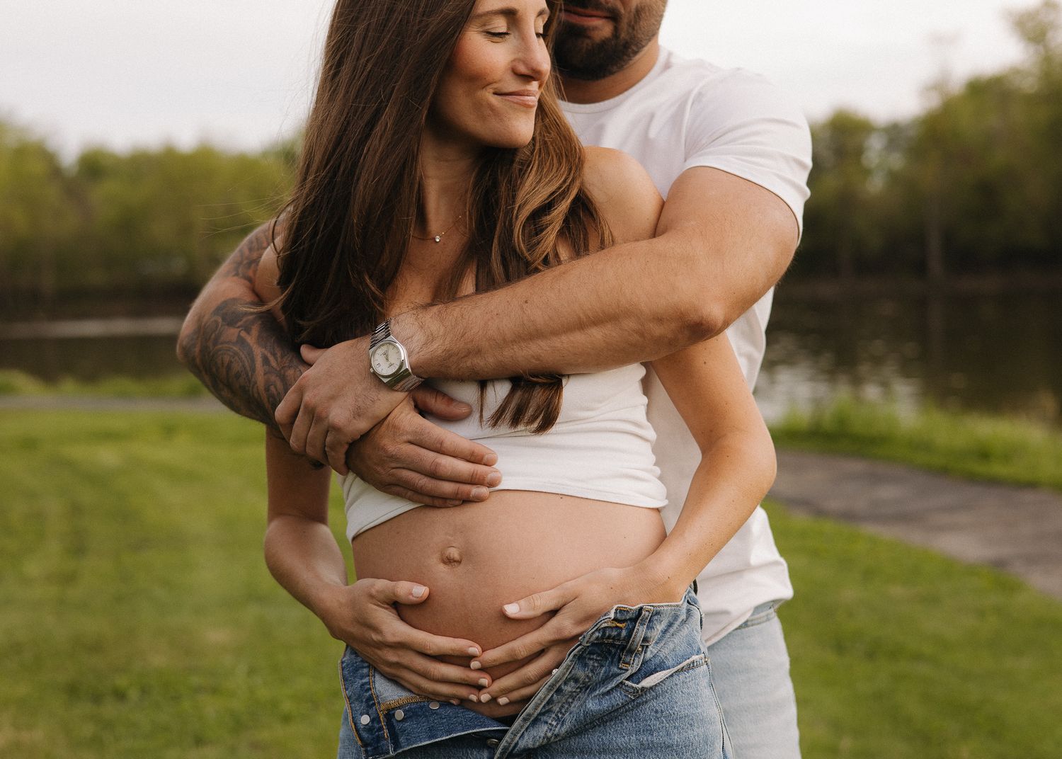 Pregnant couple outdoors near a lake, man embracing woman and cradling her baby bump.